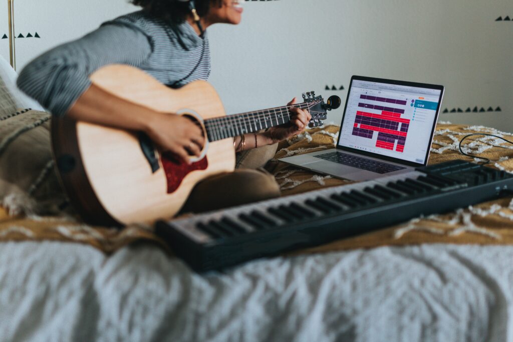 ベッドに座ってパソコンを見ながらギターを弾く男性